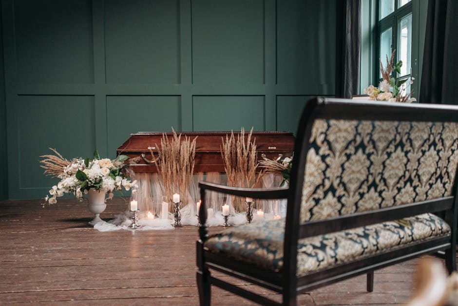 A close-up view of a floral arrangement placed on a wooden floor in front of a green paneled wall, featuring pampas grass and white flowers in a white ceramic vase. The arrangement is complemented by