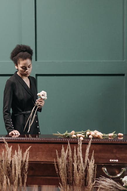 A woman with dark, curly hair and a black blazer holding a single white flower with pink accents, standing beside a wooden casket decorated with light pink roses and greenery. The arrangement is laid