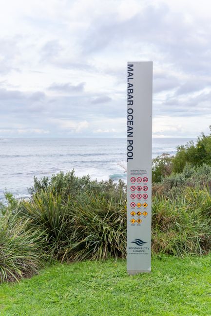 A tall, vertical signpost with a grey background displaying the words 'MALABAR OCEAN POOL' in black text, situated on a grassy area near the coast at Bromley. The sign includes various safety symbols