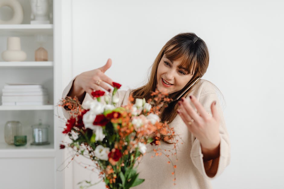 A woman with brown hair and a beige sweater is holding a vibrant floral bouquet with both hands, speaking on a mobile phone. The bouquet features a variety of fresh flowers, including white, red, and