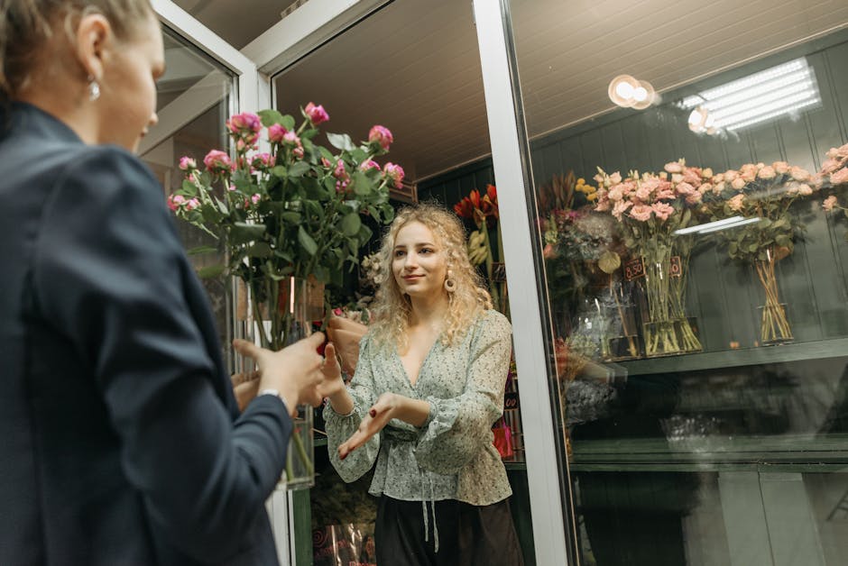 A young woman with curly blonde hair and fair skin, wearing a light floral blouse, is inside a flower shop through a glass door. She is receiving a large, vibrant bouquet of pink roses with green leav