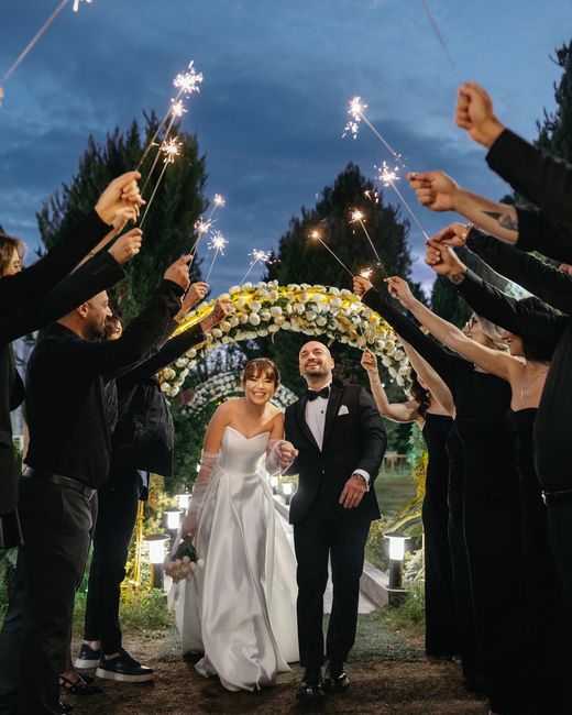A bride and groom walking hand in hand under a floral archway at night, surrounded by guests dressed in black who are holding sparklers aloft. The bride wears a strapless white wedding gown with a ful