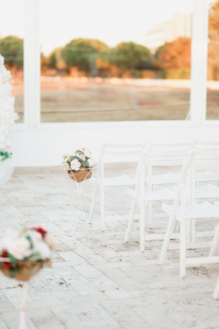 A floral arrangement featuring a small bouquet of white roses, pink peonies, and greenery, placed in a simple light brown paper wrapping with a white ribbon. The bouquet is positioned on a delicate wh