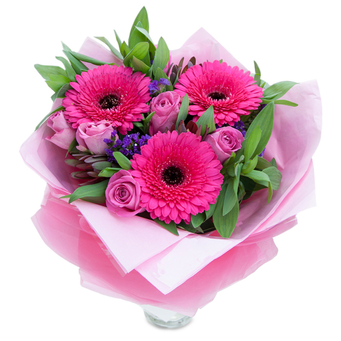 I picture this bouquet as a playful yet graceful tribute to pink's many moods, arranged and wrapped with the kind of hands-on attention we practise in our Bickley studio. Three wide-faced Gerbera daisies in saturated fuchsia open like cheerful suns, each with a dark, almost black eye that contrasts beautifully with the vivid petals. Around them, roses in a soft lavender-pink or dusty rose hue sit with petals tightly furled, their velvety surfaces suggesting a delicate perfume that mingles with the green, slightly resinous scent of eucalyptus and salal. Scattered pockets of small purple statice peek through the composition, adding a whisper of wildflower texture and a cool, blue-tinged note amid the warm pinks. The blooms rest on a foundation of leafy greenery that gives the bouquet a full, garden-fresh silhouette; every leaf is placed to complement the flowers' shapes and to catch a hint of morning light. The wrap is layered - pale pink matte tissue closest to the stems and a translucent, crinkled fuchsia-pink cellophane outside - folded with tidy, artisan folds that shine faintly and make opening the bouquet feel like unwrapping a small celebration. A clear glass vase rim is visible at the base, ready to be filled and set on a High Street table in Bickley or a kitchen shelf in nearby Chislehurst. This arrangement carries optimism and warmth, suited for birthdays, romantic notes, or to lift someone's spirits with a bright, thoughtful gesture.